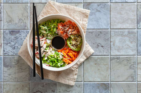 Pan Asian trendy Homemade Poke Bowl with Salmon, Rice, edamame, fresh vegetables and soy sauce, served with chopsticks on ceramic tile background.の写真素材