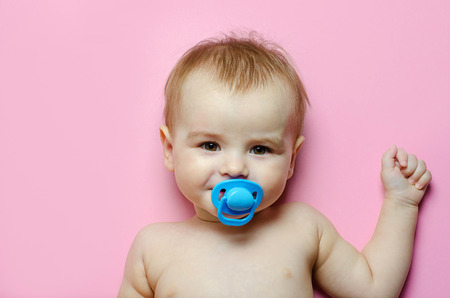 Cheerful, happy kid boy with blue pacifier lying on pink background.