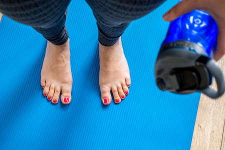 Woman practicing yoga, holding a bottle of water. Feet with pink pedicure of woman on blue mat, first person top view. Concept of indoor workout, exerciseの写真素材