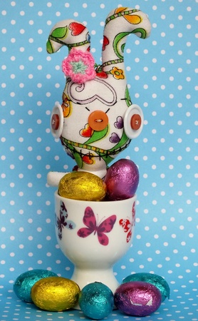 Unusual easter bunny in egg cup with small chocolate foil wrapped easter eggs on a blue spotty background の写真素材