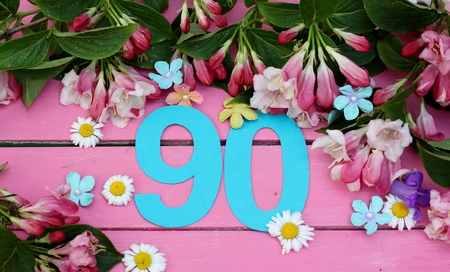 Number 90 in turquiose paper cut , surronded by spring flowers of daisies, blue bells, blossom and leaves on a bright pink painted wooden floor , image for birthday or anniversary card or invitationsの写真素材