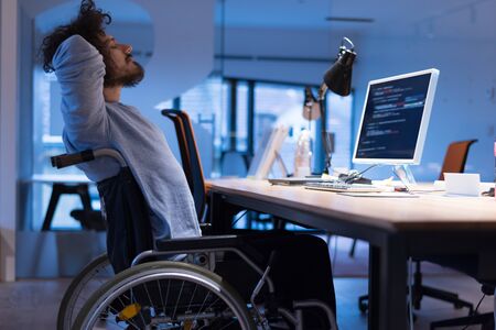 Disabled web developer in the wheelchair works in the office at the computer while performing in...