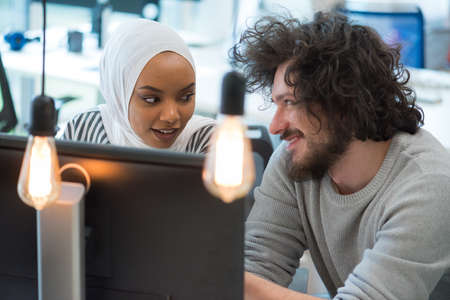 Smiling african muslim business woman talking to male colleague at work sharing office desk with...