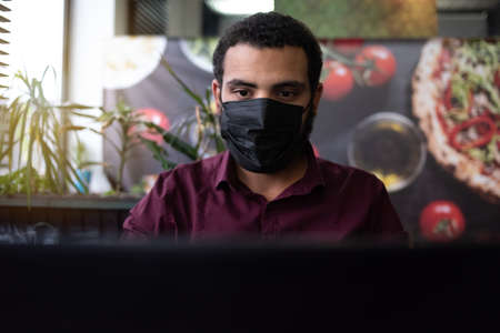 African businessman or developer wearing surgical face mask in the restaurant and typing on laptop -...