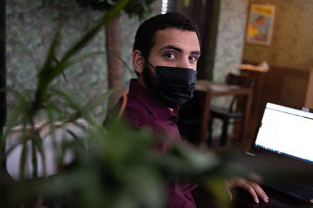 African businessman or developer wearing surgical face mask in the restaurant and typing on laptop -...