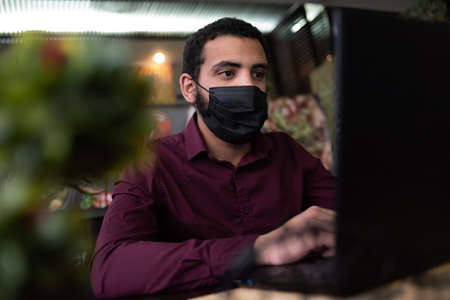 African businessman or developer wearing surgical face mask in the restaurant and typing on laptop -...