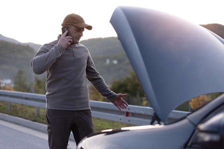 Angry young man waiting a help while sitting near the broken car at the side of road. Roadside...