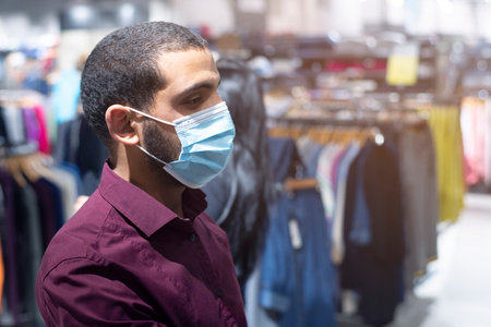 Elegant african businessman wearing coronavirus protective mask looking for suit inside man's chlothes shop or boutique at modern shopping mallの写真素材