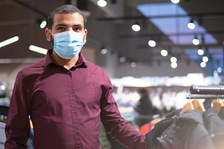 Elegant african businessman wearing coronavirus protective mask looking for suit inside man's chlothes shop or boutique at modern shopping mallの写真素材