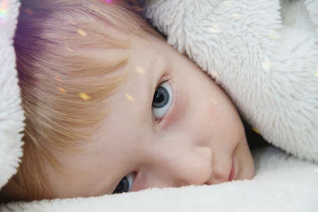 Cozy holidays at home. Cute little child lying under white knitted blanket and playingの写真素材