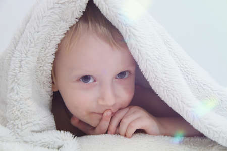 Cozy holidays at home. Cute little child lying under white knitted blanket and playingの写真素材