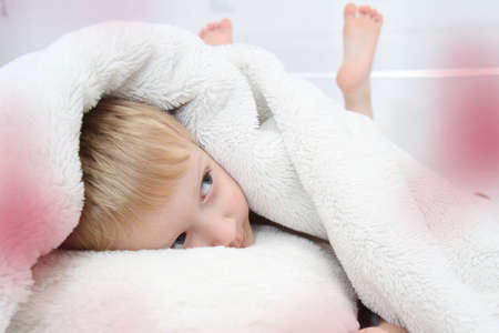 Cozy holidays at home. Cute little child lying under white knitted blanket and playing