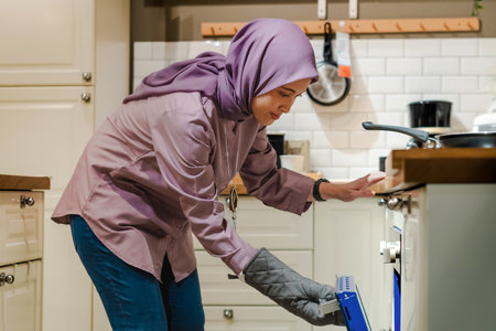 Beautiful Asian woman in hijab cooking in the kitchen, using oven and cooking glovesの写真素材