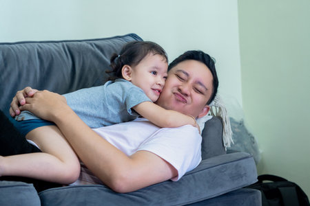 Asian man lying on sofa in living room with little girl on top, both smiling, father and daughter...