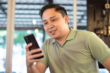 Indonesian man sitting in indoor cafe, looking at cellphone or chatting on phone with a smile,...