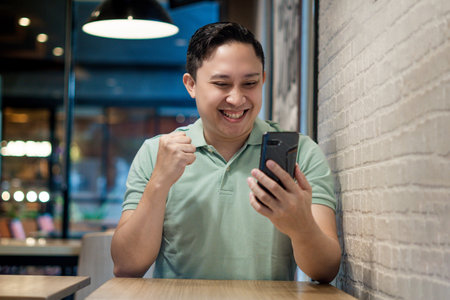 Asian man Engaging with Smartphone in a Modern Cafe Setting. Looking to good news on his phone...