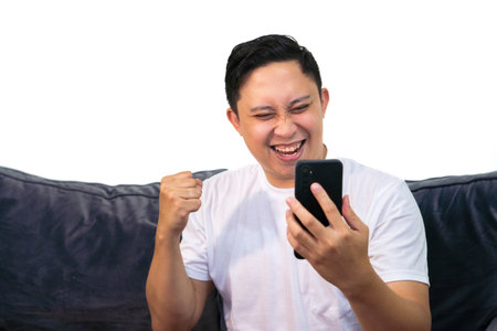 Adult Asian man in white t-shirt sitting on sofa, holding cellphone with winning gesture.の写真素材