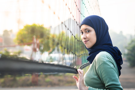 Muslim woman in hijab with brown eyes holding volleyball net in morning sunlight.
