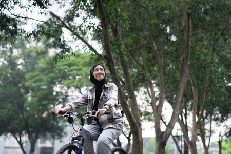 Young cheerful hijab woman, wearing casual flannel riding bicycle bike on sidewalk in city enjoying being outdoors, look aside. People active urban healthy lifestyle cycling concept.の写真素材