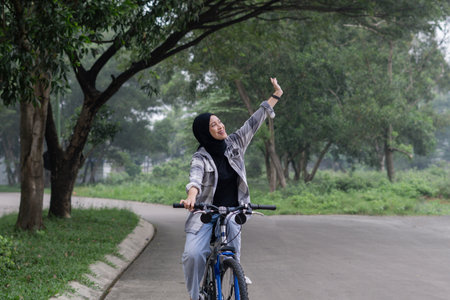 Young cheerful hijab woman, wearing casual flannel riding bicycle bike on sidewalk in city enjoying...