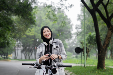 Young cheerful hijab woman, wearing casual flannel riding bicycle bike on sidewalk in the city park...