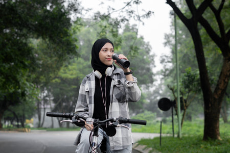 Young cheerful hijab woman, wearing casual flannel riding bicycle bike on sidewalk in the city park...