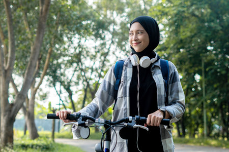 Young cheerful hijab woman, wearing casual flannel riding bicycle bike on sidewalk in the city park enjoying being outdoors, look aside or ahead. People active urban healthy lifestyle cycling concept.の写真素材