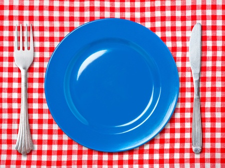Empty blue dinner plate with fork and knife on red and white checked tablecloth.
