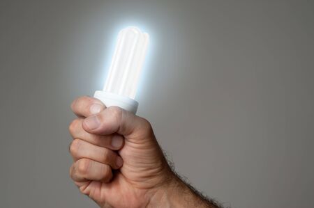Hand holding a glowing light bulb on gray background.