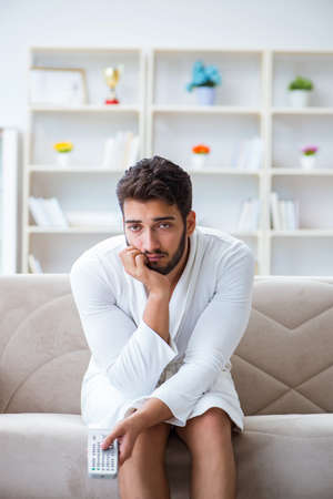 Young man in a bathrobe watching television at home on a sofa coの写真素材