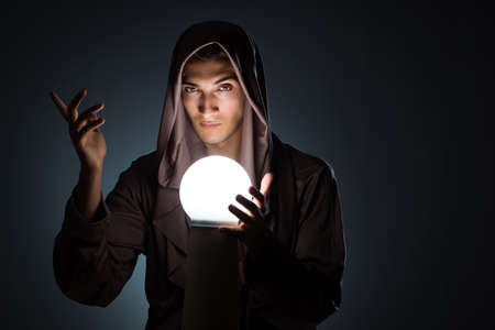 Young wizard with crystal ball in dark room