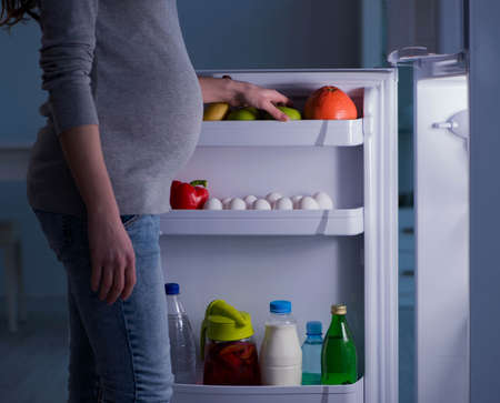 The pregnant woman near fridge looking for food and snacks at nightの写真素材