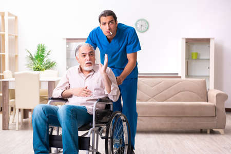 Old man in wheel-chair and young bad caregiver indoorsの写真素材