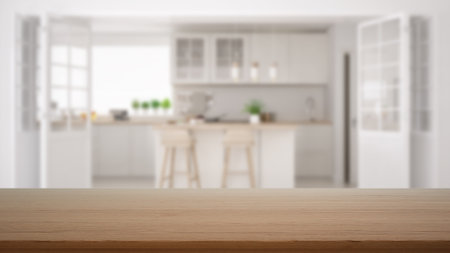 Empty wooden table, desk or shelf with blurred view of scandinavian white kitchen with cabinets, island and stools, decors and accessories, modern interior design conceptの写真素材
