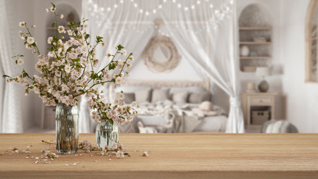 Wooden table, desk or shelf close up with branches of cherry blossoms in glass vase over blurred view of bohemian bedroom with canopy bed, boho interior design conceptの写真素材