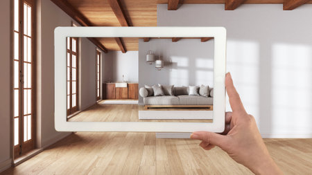 Augmented reality concept. Hand holding tablet with AR application used to simulate furniture and design products in empty wooden interior with parquet, living room and kitchenの写真素材