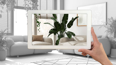 Augmented reality concept. Hand holding tablet with AR application used to simulate furniture and design products in total white background, urban jungle living roomの写真素材