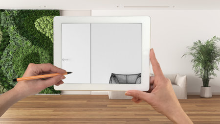 Hands holding and drawing on tablet showing sitting waiting room with vertical garden, details CAD sketch. Real finished interior in the background, architecture design presentationの写真素材