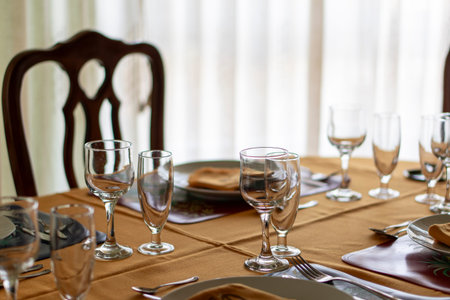 A table with a chair and a tablecloth with a few glasses and utensils. The table is set for a meal