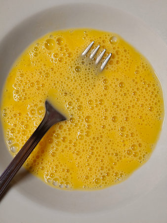 Whisked raw eggs in a white bowl showing foamy, bubbly yellow texture with a metal fork for beating fresh, homemade breakfast or baking preparation, close up top viewの写真素材