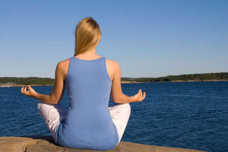 a young blond woman sits on the stones with the ocean in front of her doing some yoga and recreatingの写真素材