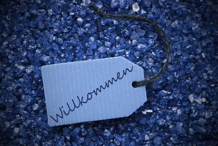 One Blue Label Or Tag With Black Ribbon On Blue And Purple Small Stones As Background With German Text Willkommen Means Welcome With Frameの写真素材