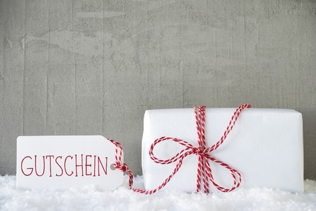 One Christmas Gift Or Present On Snow. Cement Or Concrete Wall As Background. Modern And Urban Style. Card For Birthday Or Seasons Greetings. Label With German Text Gutschein Means Voucherの写真素材