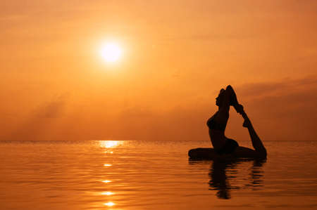 Silhouette young woman practicing yoga on the beach at sunsetの写真素材