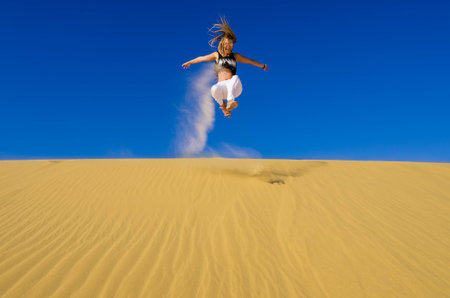 girl jumping on the yellow sand dune. Clear blue sky in the background.