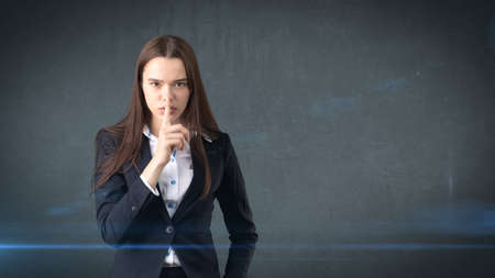 Young businesswoman portrait in suit standing and finger up near her lips with hush sign, background with copyspace.の写真素材