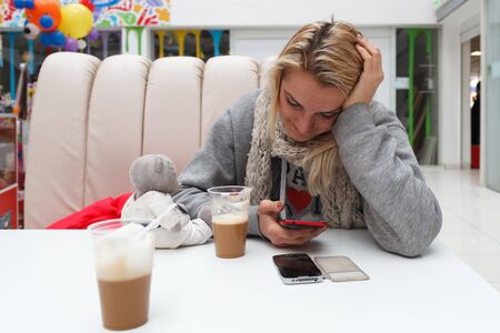 Girls in a jacket sits at a table with coffee and holds phone in hand