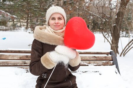 The girl in winter clothes keeps the red balloon in the form of heart in the park in the winter in the middle of snowの写真素材