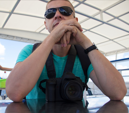 The young fair-haired man in a t-shirt, a backpack and sunglasses sits at a table against the background of the cruise ship of the liner in the summer の写真素材
