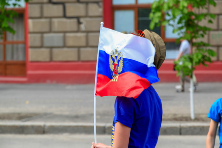 VOLGOGRAD, RUSSIA - May 9, 2018: The boy in a green garrison cap flies a flag of Russia with the image of the coat of arms with a two-headed eagleのeditorial素材